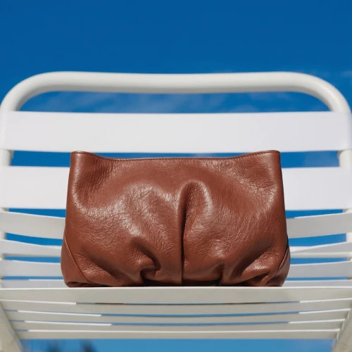 Clutch Inanna din piele naturala maro tabac, asezat pe un scaun alb, pe fundal albastru